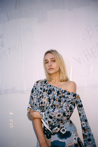 Portrait of a blonde woman in floral top and skirt, posing indoors against white wall with handwritten text and studio lighting.