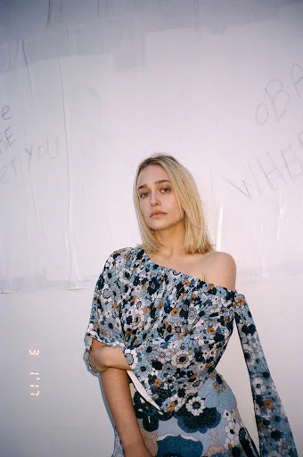 Portrait of a blonde woman in floral top and skirt, posing indoors against white wall with handwritten text and studio lighting.