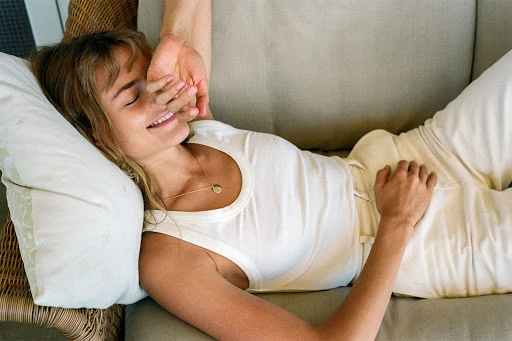 Woman with bangs wearing a white tank top lying on a beige sofa, smiling while covering her eyes with her hand.