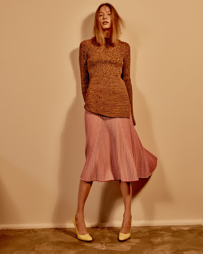 A woman in a brown textured sweater and pink patterned midi skirt poses against a beige wall, completing the look with yellow heels.