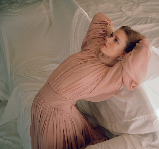 Woman in a pink pleated dress leaning backward among white draped fabric with her hands behind her head.