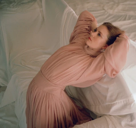 Woman in a pink pleated dress leaning backward among white draped fabric with her hands behind her head.
