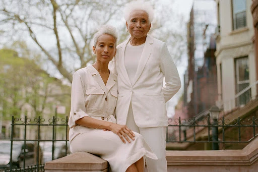 Two elegant women, one seated and one standing, pose in coordinated neutral outfits against an urban backdrop with greenery and brownstones.