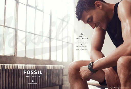 A tanned man sits in a fitness environment, wearing a smartwatch, showcasing sportswear and a healthy lifestyle against a bright background.