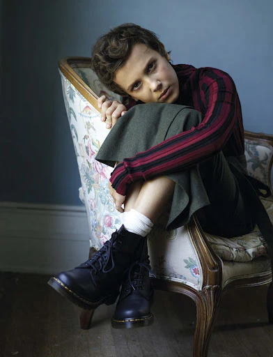 Young adult with short curly hair sits on a vintage chair wearing black boots, a striped shirt, and a skirt in an indoor setting.