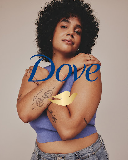 Confident woman with natural curly hair poses in front of Dove logo in a studio setting, showcasing body positivity and self-love.