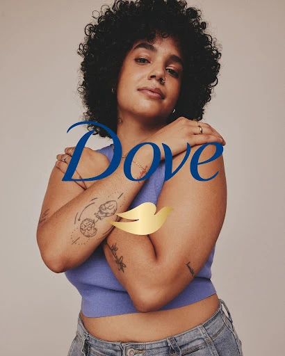 Confident woman with natural curly hair poses in front of Dove logo in a studio setting, showcasing body positivity and self-love.