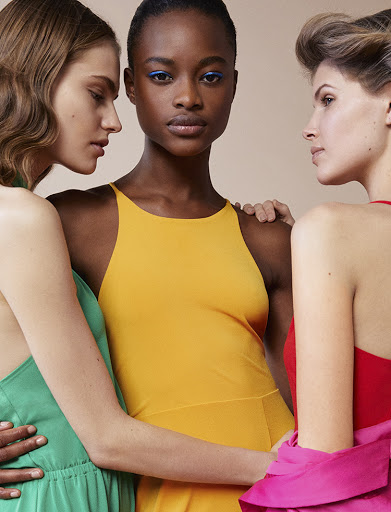 Three women in a close studio shot. The woman in the middle wears a yellow dress with blue eye makeup, flanked by two women in colorful dresses.