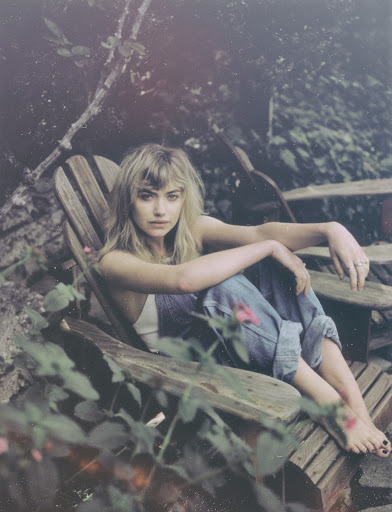 Young blonde woman in overalls relaxing on wooden Adirondack chair outside with leafy green background. Barefoot, casual look, natural light.