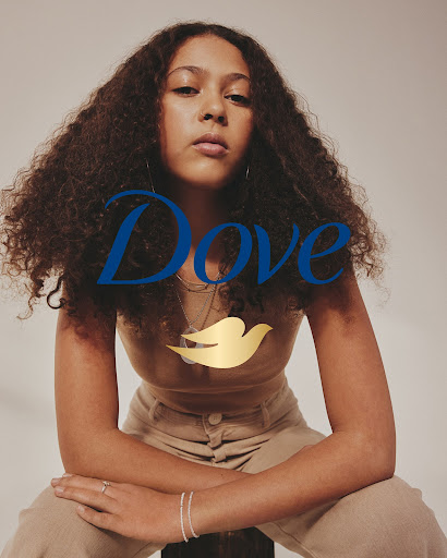 Portrait of a young adult woman with curly brown hair, wearing a beige top and pants, seated against a neutral background, with Dove logo.