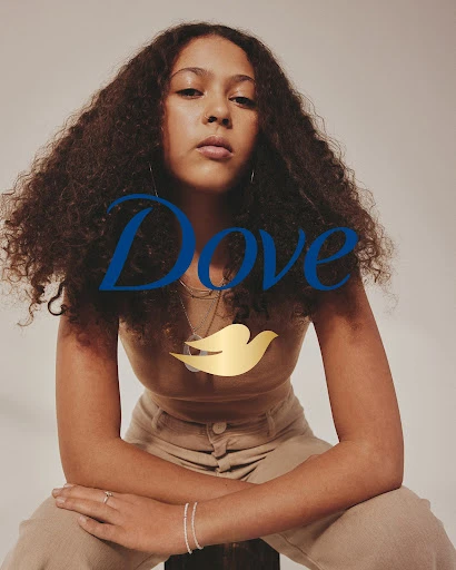 Portrait of a young adult woman with curly brown hair, wearing a beige top and pants, seated against a neutral background, with Dove logo.