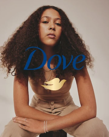 Portrait of a young adult woman with curly brown hair, wearing a beige top and pants, seated against a neutral background, with Dove logo.