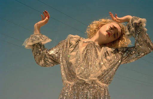Woman with blonde curly hair, eyes closed, wearing a sparkly silver dress with frills, posing against a blue sky with powerlines.
