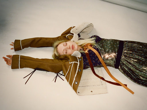 A fair-skinned young woman lies on her back on a white surface in a fashion pose, wearing a metallic dress and jacket with tan trim.