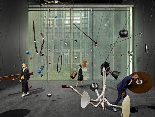 Art gallery scene featuring three people viewing a complex overhead kinetic sculpture with hanging metal and plastic pieces in an art exhibit.