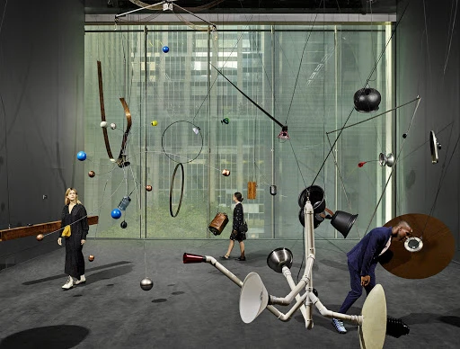 Art gallery scene featuring three people viewing a complex overhead kinetic sculpture with hanging metal and plastic pieces in an art exhibit.