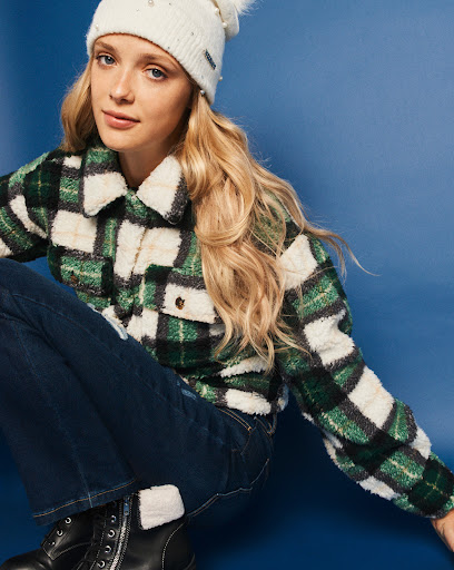 Blonde model wearing a green and white plaid jacket, dark jeans, and black boots in a studio setting on a blue backdrop.