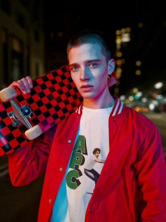 Young man with a buzzcut wearing a red jacket holding a checkered skateboard at night