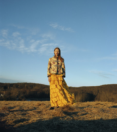 Woman in a floral jacket and yellow ruffled skirt stands in a field under a blue sky with scattered clouds and distant trees.