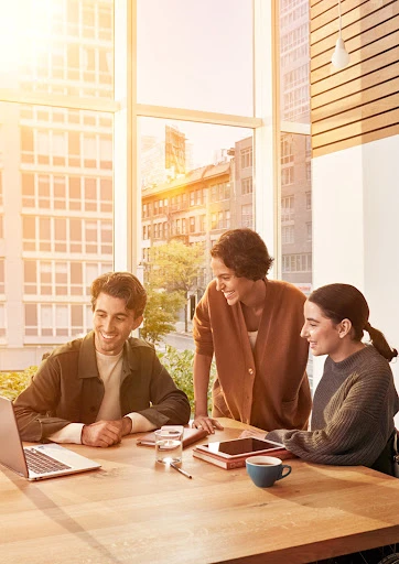 Three smiling adults collaborate at a desk in a bright, modern office. Laptop, tablet, coffee cup, and large window with cityscape.