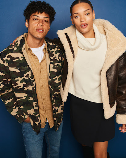 Two models pose in a studio setting with a blue background. One wears a camo jacket, the other a cream sweater and brown aviator jacket.