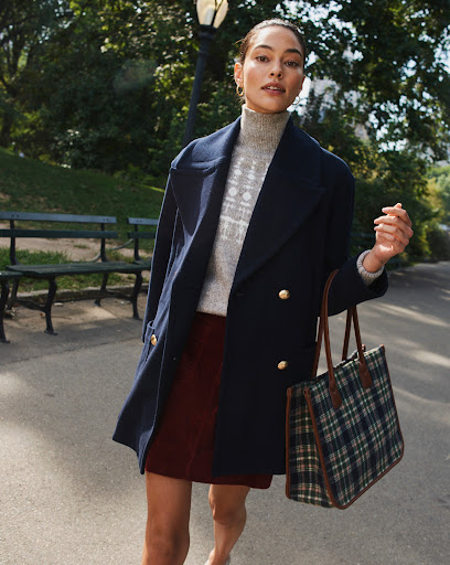 Woman in a navy pea coat and patterned turtleneck holding a plaid tote bag along a park path