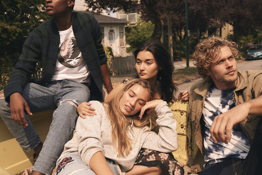 Group of four young adults in casual summer clothing sit together in an outdoor, urban environment; natural lighting, medium shot.