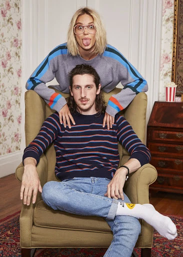 Two adults in an indoor environment: A woman stands behind an armchair with a seated man, both wearing sweaters and jeans.
