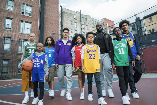 Group of diverse people in basketball jerseys standing on an outdoor court with brick buildings in the background, wearing sneakers.