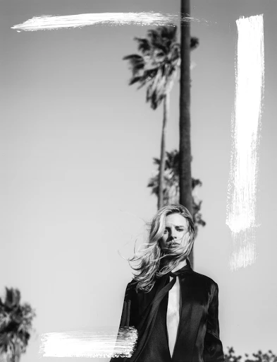 Black and white portrait of a woman with long blonde hair in a silky shirt, posing outdoors near palm trees under a bright sky.