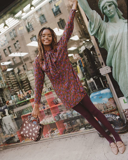 A young Black woman smiles in a floral dress next to a Statue of Liberty figure, holding a purse in an urban setting.