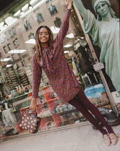 A young Black woman smiles in a floral dress next to a Statue of Liberty figure, holding a purse in an urban setting.