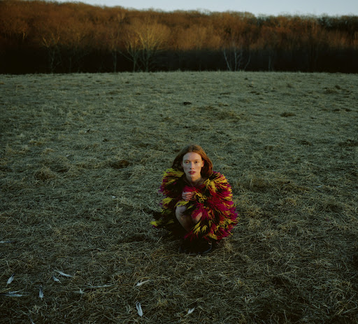 A young adult woman crouches in a grassy field wearing a colorful feathered garment under a partly cloudy sky with wooded hills.