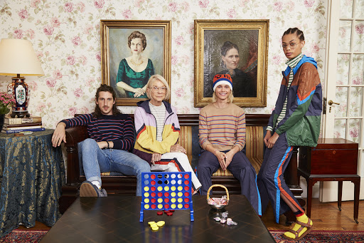 Four people sit on a couch in an interior with floral wallpaper and paintings. A Connect Four game sits on a coffee table.