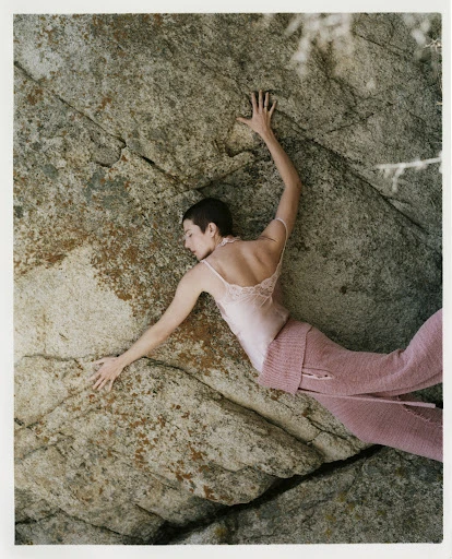 Woman in a pink top and trousers lying on a large rock, outdoors. She is holding onto the rock with her hands above her head.