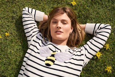 Woman lying on green grass wearing a white sweater with black stripes and a bee motif, styled by Liz Mcclean.