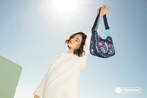 Woman in white top holding up a patterned blue shoulder bag against a bright blue sky with direct sunlight.