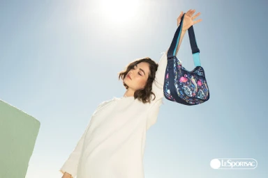 Woman in white top holding up a patterned blue shoulder bag against a bright blue sky with direct sunlight.