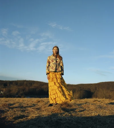 Model standing in a dry field wearing a patterned jacket and a bright yellow ruffled skirt under a blue sky.