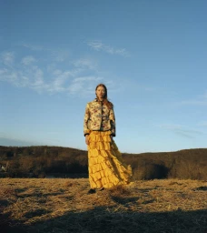 Model standing in a dry field wearing a patterned jacket and a bright yellow ruffled skirt under a blue sky.