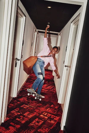 A smiling woman wearing a pink shirt and blue jeans rides a skateboard down a hotel hallway with a large brown bag.