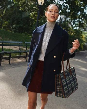 Woman in a navy pea coat and patterned turtleneck holding a plaid tote bag along a park path
