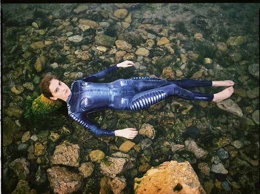 Woman in a full body suit lying in shallow water among rocks, looking up at camera. Outdoor shot with natural light. 