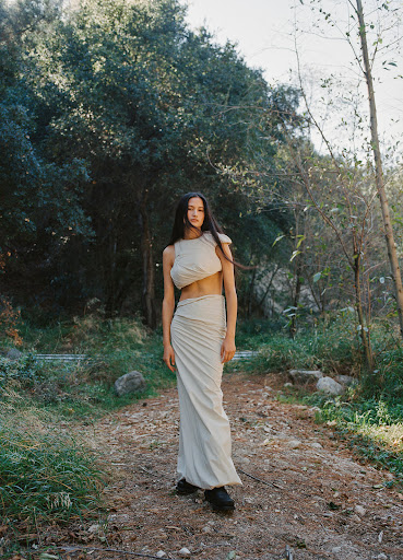 A slender woman with long hair poses on a dirt path wearing a two-piece tan outfit, surrounded by a forest with dappled sunlight.