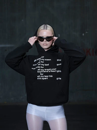 Woman in black hoodie and sunglasses poses against a dark wall. The black hoodie has white text on it. She is wearing white shorts.
