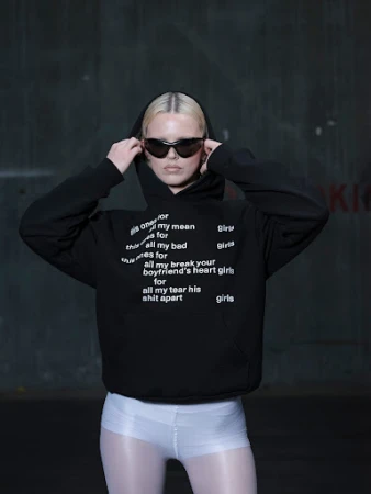 Woman in black hoodie and sunglasses poses against a dark wall. The black hoodie has white text on it. She is wearing white shorts.