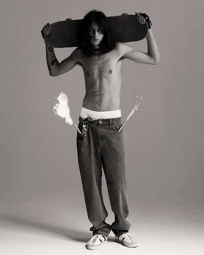 Full-length portrait of a topless male skater holding a skateboard behind his head, wearing jeans, and sneakers in a photo studio.