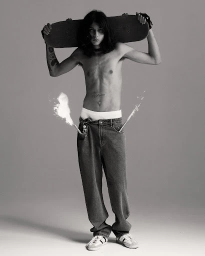 Full-length portrait of a topless male skater holding a skateboard behind his head, wearing jeans, and sneakers in a photo studio.