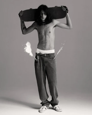 Full-length portrait of a topless male skater holding a skateboard behind his head, wearing jeans, and sneakers in a photo studio.