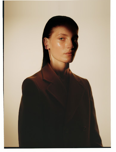 Portrait of a young adult female with dark hair, wearing an oversized brown suit jacket in a studio setting with diffused lighting.